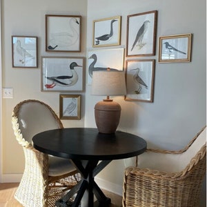 May include: A black round table with a wood top and a black X-shaped base sits in a corner of a room. Two wicker chairs with white cushions are placed on either side of the table. The wall behind the table is decorated with framed prints of birds.