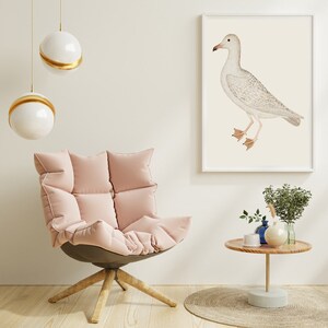 May include: A pink, plush armchair with a white, round, gold-rimmed pendant light hanging above it. A framed print of a white seagull on a cream background hangs on the wall behind the chair. A small wooden side table with a blue vase and a green plant sits in front of the chair.