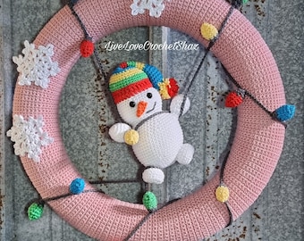 May include: A pink crocheted wreath with a snowman hanging from it. The wreath is decorated with white crocheted snowflakes and a string of colourful crocheted lights.