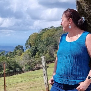 May include: A woman wearing a blue crocheted tank top with a lacy trim, standing in front of a green field and a forest.