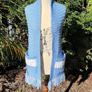May include: A light blue crocheted scarf with white accents and fringe. The scarf has two pockets on the front.