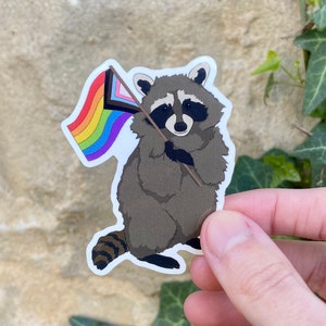 May include: A sticker of a raccoon holding a rainbow pride flag. The raccoon is brown and gray with a black nose and eyes. The flag is a rainbow with a black triangle on the top left corner.