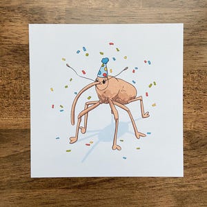 May include: A whimsical illustration on a white card features a cartoon insect wearing a blue party hat. Colorful confetti surrounds the insect. The insect has long antennae and multiple legs. The artwork is set against a wooden background.