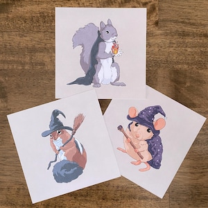 Könnte beinhalten: Drei quadratische Kunstdrucke mit skurrilen Illustrationen von Waldtieren in Hexenkostümen. Ein Eichhörnchen hält eine Flamme, ein Chipmunk hat einen Besen und eine Maus hält einen Stab. Die Drucke haben einen hellen Hintergrund.