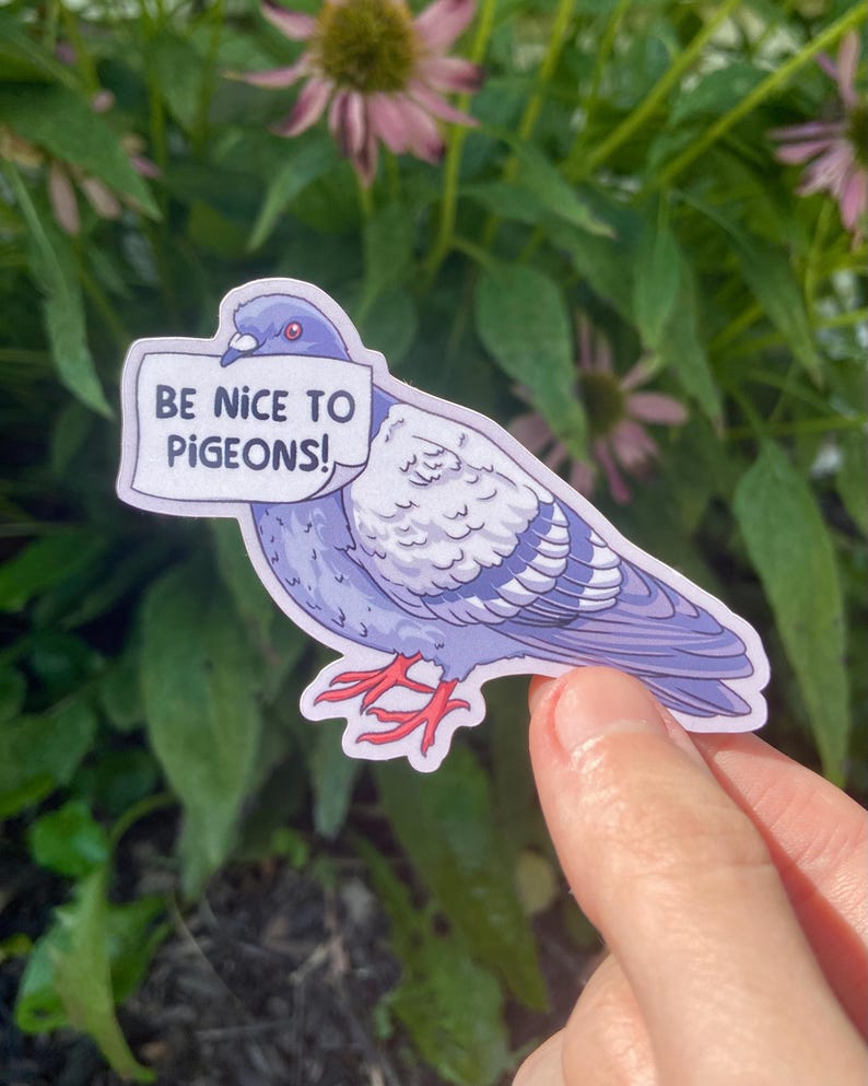 K&ouml;nnte beinhalten: Ein gestanzter Aufkleber, der eine violette und graue Taube mit roten F&uuml;&szlig;en zeigt. Die Taube h&auml;lt ein wei&szlig;es Schild im Schnabel, auf dem in schwarzen Gro&szlig;buchstaben BE NICE TO PIGEONS! steht. Die Illustration hat einen wei&szlig;en Rand.