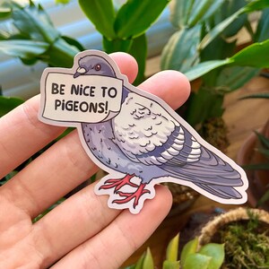 K&ouml;nnte beinhalten: Ein gestanzter Aufkleber mit einer Cartoon-Taube mit grauem und wei&szlig;em Gefieder und roten F&uuml;&szlig;en. Die Taube h&auml;lt ein wei&szlig;es Schild im Schnabel, auf dem in schwarzen Gro&szlig;buchstaben BE NICE TO PIGEONS! zu lesen ist.