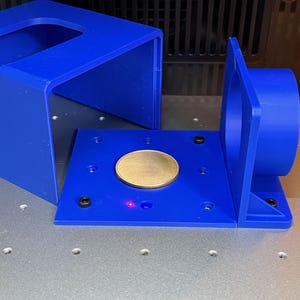 May include: A blue 3D-printed plastic housing component featuring a flat square base with a circular cutout, a vertical side panel with a cylindrical opening, and a separate U-shaped cover. A gold-colored coin sits on the base near a red laser dot.
