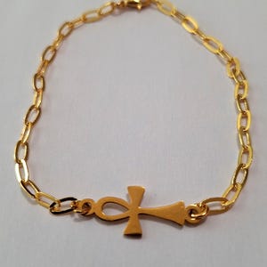 May include: Gold-colored bracelet featuring an Ankh symbol. The bracelet has a delicate chain design with an oval link pattern. The Ankh symbol is centered on the bracelet. The jewelry is displayed on a white surface.