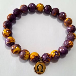 May include: A beaded bracelet featuring a mix of purple and yellow-orange round beads. The bracelet has a gold-colored charm with the Greek letter Omega. The beads have a glossy finish and are strung together to form a complete circle.