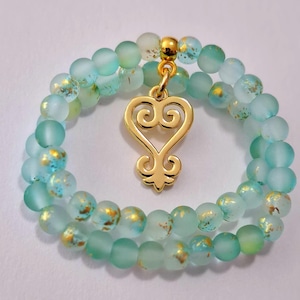 May include: A double-strand bracelet composed of frosted light blue beads with gold-colored flecks. The bracelet features a central gold-colored metal charm shaped like a stylized heart with symmetrical swirling patterns at the top and bottom.