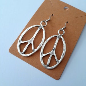 May include: A pair of silver peace sign earrings on a brown card. The earrings are oval shaped and have a textured surface.