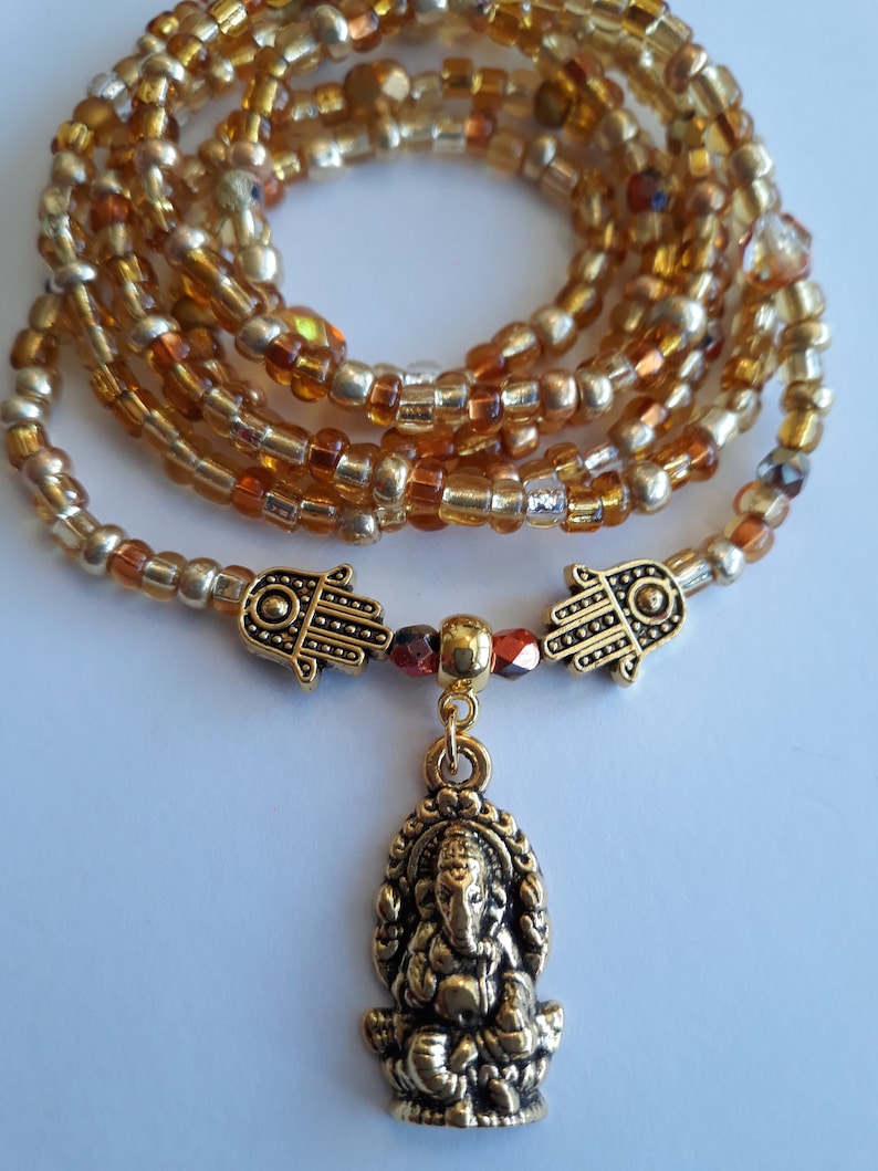 May include: A gold beaded wrap bracelet with a gold charm of the Hindu deity Ganesha. The bracelet is made with small, round beads in shades of gold and amber.
