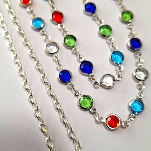 May include: Three silver chains with colorful round gemstones. The chains are layered on a white background. The gemstones are red, blue, green, and clear.