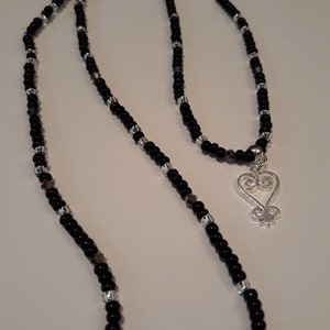 May include: A black and silver beaded necklace with a silver heart pendant. The heart pendant has a traditional African Adinkra symbol.