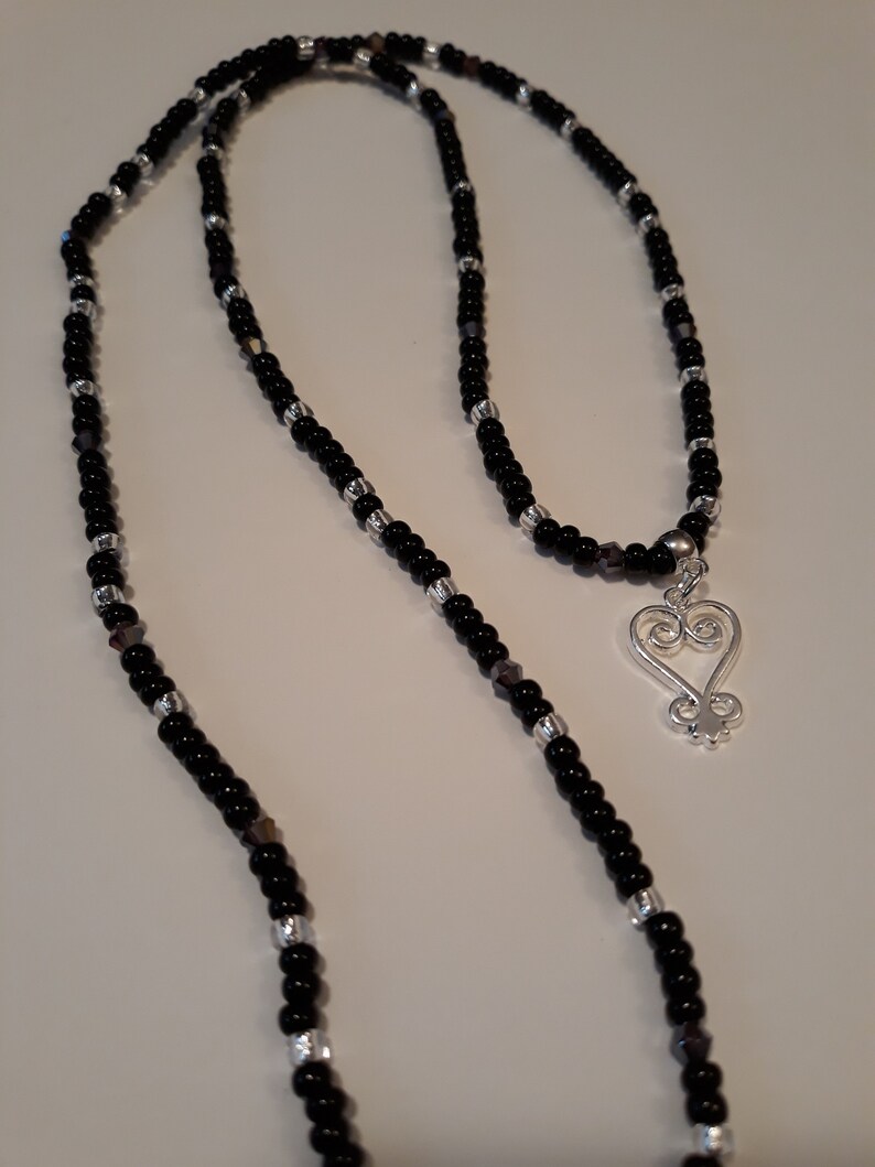 May include: A black and silver beaded necklace with a silver heart pendant. The heart pendant has a traditional African Adinkra symbol.