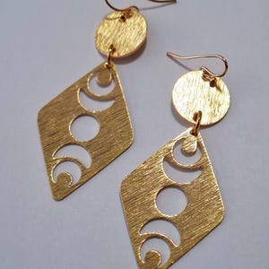 May include: Gold-colored earrings with a textured finish. Each earring features a circular top and a diamond-shaped pendant with crescent and circular cutouts. The earrings have a hook closure.