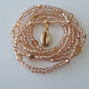 May include: A multi-strand beaded necklace with a gold-toned seashell pendant. The beads are a light peach color and have a subtle shimmer.