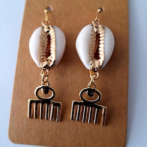 May include: A pair of gold earrings with white cowrie shells and gold comb charms. The earrings are displayed on a brown card.