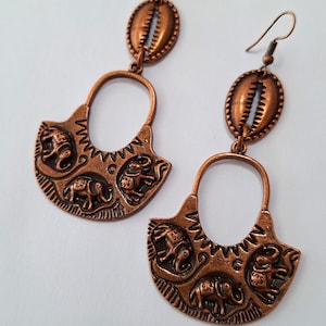 May include: A pair of copper-colored earrings. Each earring features a decorative oval top and a fan-shaped bottom with elephant designs. The earrings have a hook closure and are made of a copper-toned metal.