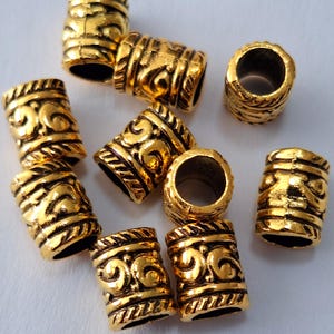 May include: A group of ten gold-colored metal beads with intricate designs. The beads are cylindrical and have a slightly textured surface.