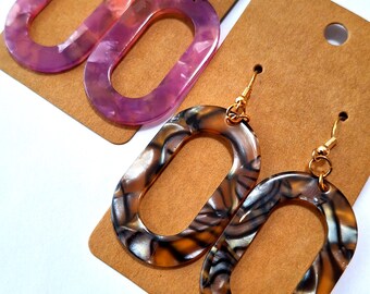 May include: Two pairs of oval-shaped dangle earrings. One pair is a marbled purple and pink, the other is a marbled brown, black, and white. Both pairs have gold-colored hooks and are displayed on brown earring cards.