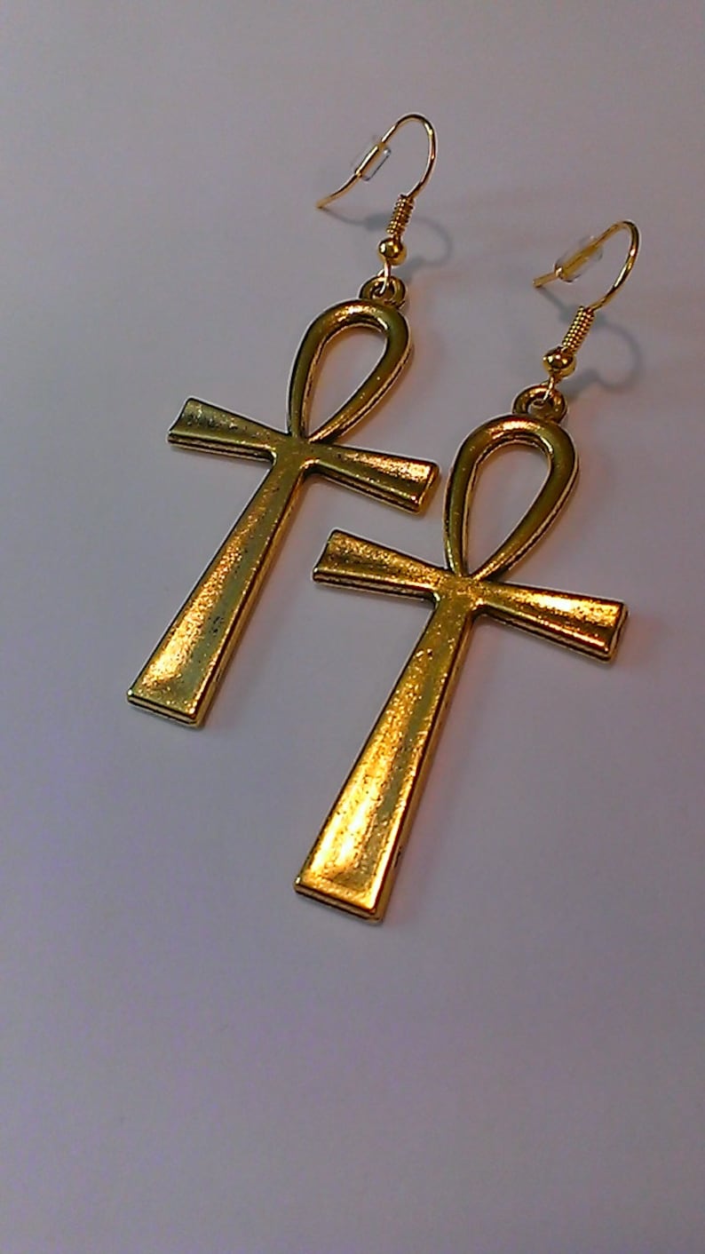 May include: A pair of gold-toned Ankh earrings with a dangling design. The earrings are made of metal and have a shiny finish.