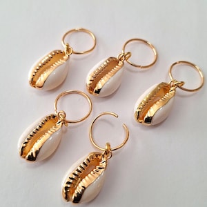 May include: Five gold-colored hair charms with white and gold seashell pendants. Each charm features a small, open ring for attachment. The seashells have a detailed, textured design.