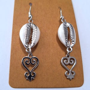 May include: A pair of dangle earrings featuring white seashells with silver-colored metal accents. Each earring has a decorative silver-colored heart-shaped charm. The earrings are displayed on a brown card.