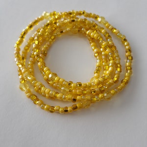 May include: A yellow beaded waist bead necklace with multiple strands of beads. The beads are small and square shaped.