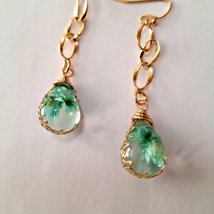 May include: A pair of gold dangle earrings with a teardrop-shaped pendant featuring a pressed flower design. The flower is green and white and is encased in clear resin.