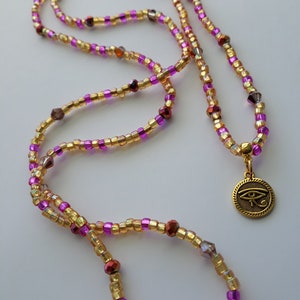 Puede incluir: Un collar largo de cuentas con cuentas doradas, moradas y rojas. El collar tiene un colgante dorado con un s&iacute;mbolo de ojo. Las cuentas peque&ntilde;as y redondas est&aacute;n sobre una superficie blanca.