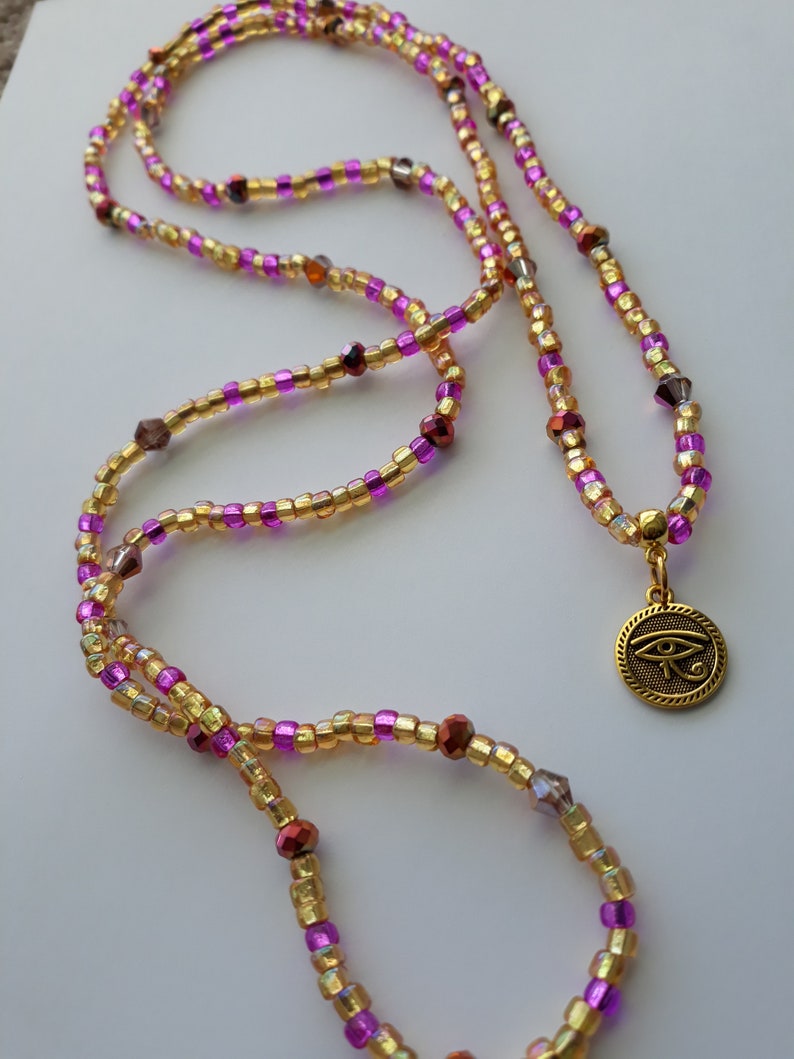 Puede incluir: Un collar largo de cuentas con cuentas doradas, moradas y rojas. El collar tiene un colgante dorado con un s&iacute;mbolo de ojo. Las cuentas peque&ntilde;as y redondas est&aacute;n sobre una superficie blanca.