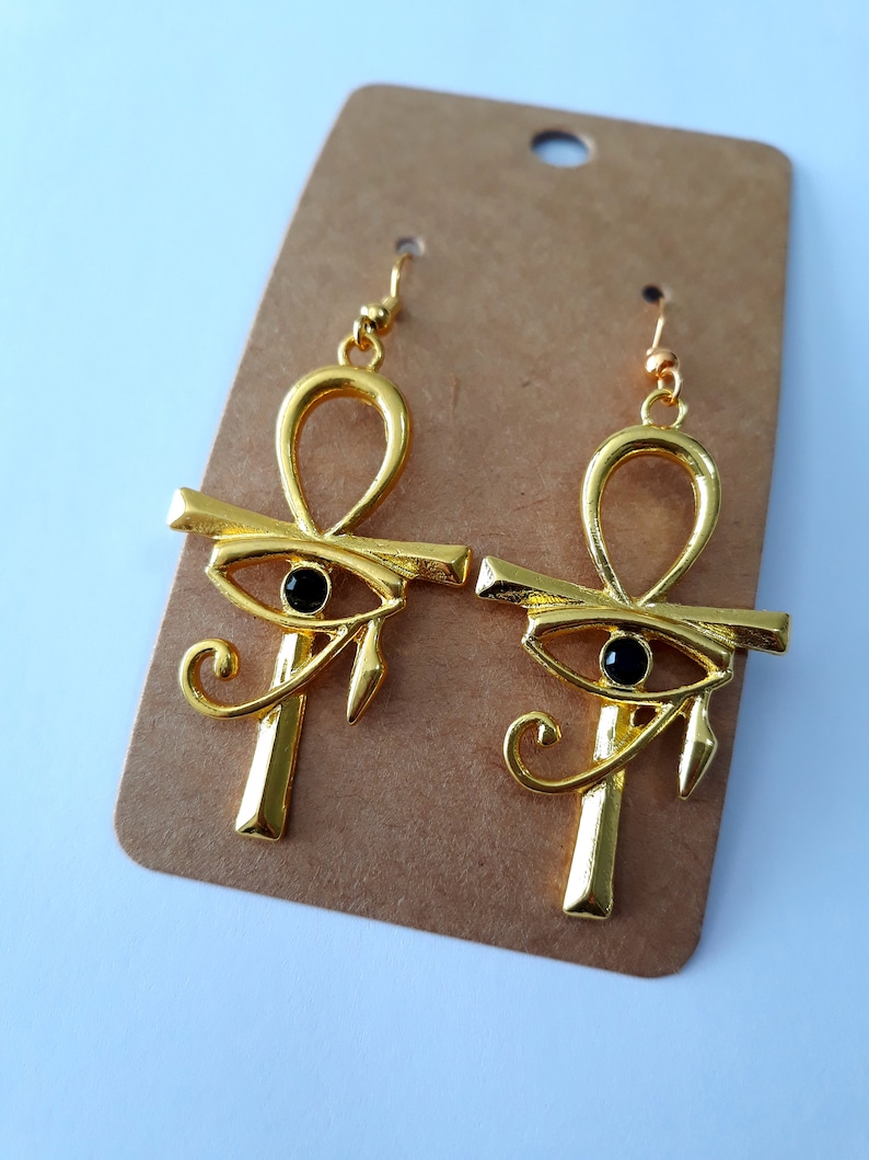 May include: A pair of gold-tone earrings with an ankh design and a black eye of Horus charm. The earrings are hanging on a brown card.