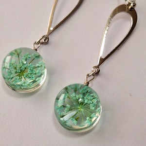 May include: Silver dangle earrings with a teardrop shaped design. Each earring features a round, clear resin charm with a pressed green flower inside.