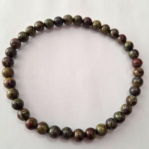 May include: A beaded bracelet composed of small, round beads in shades of green, brown, and burgundy. The bracelet is strung together, forming a complete circle. The beads have a speckled appearance, and the bracelet is set against a white background.