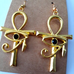May include: A pair of gold-tone earrings featuring the Egyptian Ankh symbol with a black eye of Horus charm. The earrings have a hook closure.
