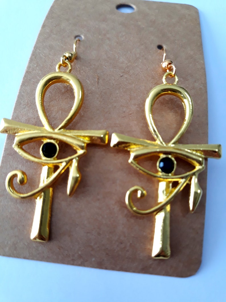 May include: A pair of gold-tone earrings featuring the Egyptian Ankh symbol with a black eye of Horus charm. The earrings have a hook closure.