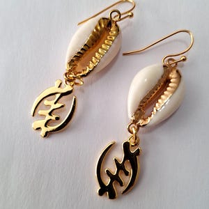 May include: Gold earrings with a white cowrie shell and a gold Adinkra symbol pendant. The symbol is the Sankofa symbol, which means "return and get it".