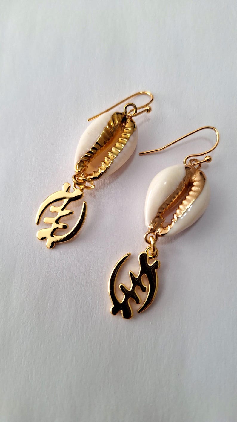 May include: Gold earrings with a white cowrie shell and a gold Adinkra symbol pendant. The symbol is the Sankofa symbol, which means "return and get it".