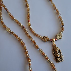 May include: A gold beaded necklace with a charm featuring a small golden figure of Ganesha and two Hamsa hands. The beads are a mix of gold, brown, and clear.
