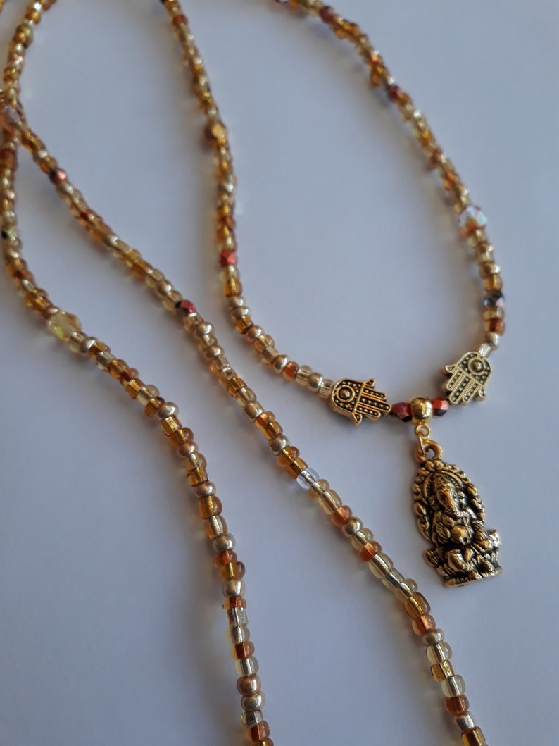 May include: A gold beaded necklace with a charm featuring a small golden figure of Ganesha and two Hamsa hands. The beads are a mix of gold, brown, and clear.