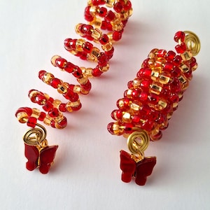May include: Two red and gold beaded hair coils with butterfly charms. The coils are made of small red and gold beads that are strung together in a spiral shape. The butterfly charms are attached to the bottom of the coils.