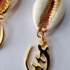 May include: Gold earrings with a white cowrie shell and a gold Adinkra symbol pendant. The symbol is the Sankofa, which means "return and get it".