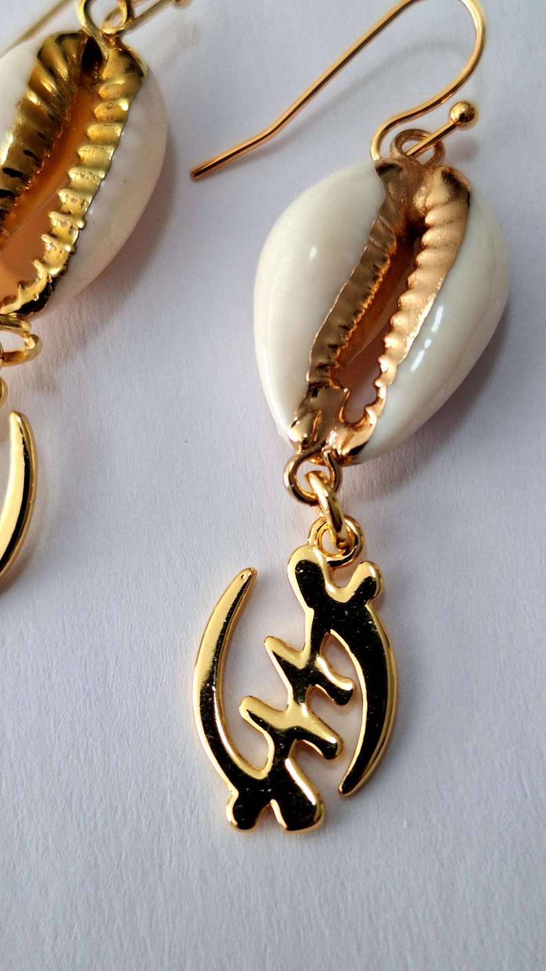 May include: Gold earrings with a white cowrie shell and a gold Adinkra symbol pendant. The symbol is the Sankofa, which means "return and get it".