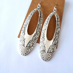 May include: A pair of silver-toned oval earrings with a decorative swirl pattern. The earrings are suspended from a hook and are displayed on a brown card against a white background. The earrings have a cutout center.