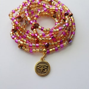 Puede incluir: Una cadena de cintura de cuentas en tonos dorados, rosas y morados. La cadena presenta peque&ntilde;as cuentas coloridas y un dije dorado con un dise&ntilde;o del Ojo de Horus. La cadena est&aacute; enrollada y descansa sobre una superficie blanca.