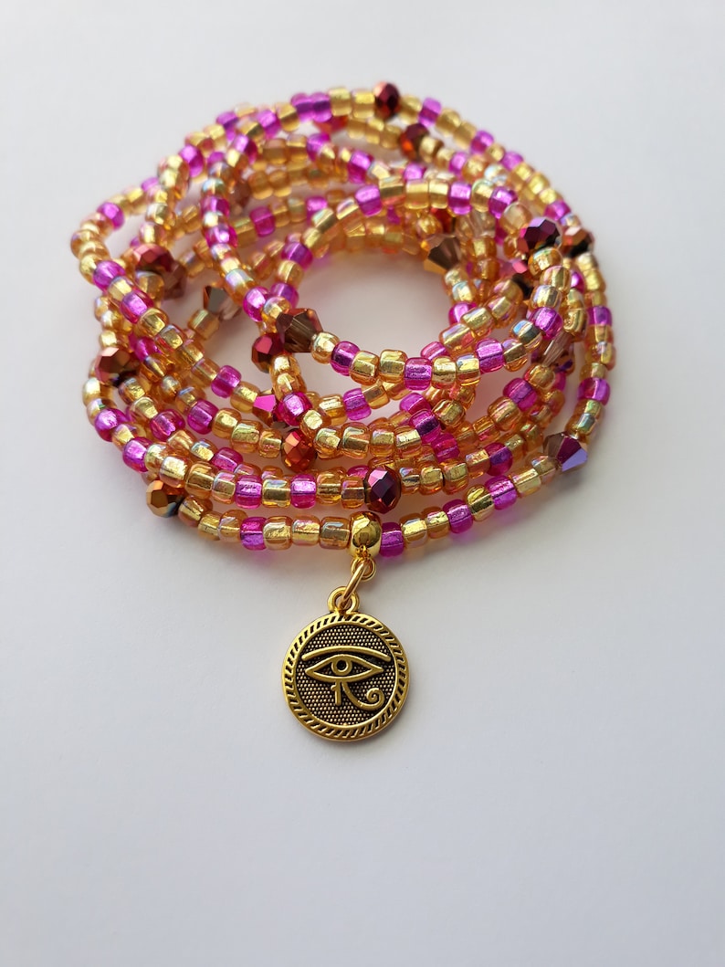 Puede incluir: Una cadena de cintura de cuentas en tonos dorados, rosas y morados. La cadena presenta peque&ntilde;as cuentas coloridas y un dije dorado con un dise&ntilde;o del Ojo de Horus. La cadena est&aacute; enrollada y descansa sobre una superficie blanca.