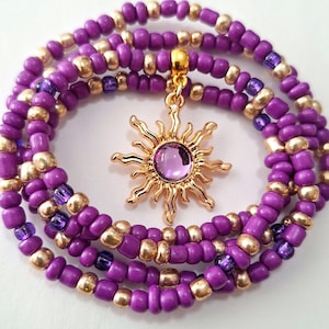 May include: A layered beaded necklace featuring purple and gold beads. A gold-colored sun pendant with a purple center stone hangs from the necklace. The necklace is displayed on a white background.