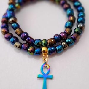 May include: A layered beaded necklace with a metallic, iridescent finish. The beads transition through shades of blue, purple, and gold. A small, colorful ankh pendant hangs from the necklace, adding a touch of symbolism. The necklace is displayed against a white background.