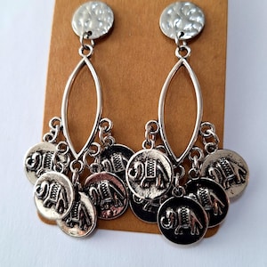 May include: Silver-toned dangle earrings with a bohemian design. Each earring features a textured disc stud, an elongated oval center, and multiple dangling coin charms with elephant motifs. The earrings are displayed on a brown card.